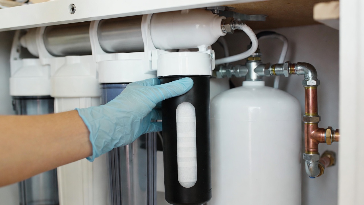 Hand wearing a blue glove replacing a black sediment filter cartridge in an under-sink reverse osmosis system